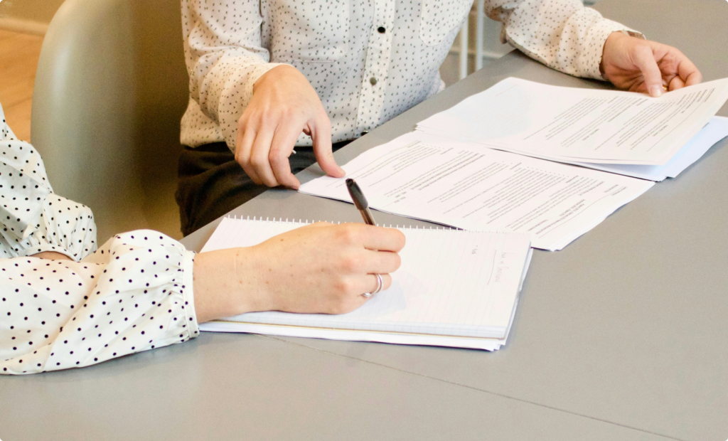 Two professionals in a business meeting reviewing and discussing documents, with one taking notes, symbolizing teamwork, negotiation, and strategic planning.