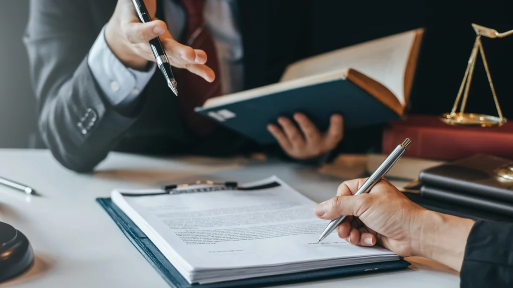 a person signing documents while another one holds a large book and talks to them
