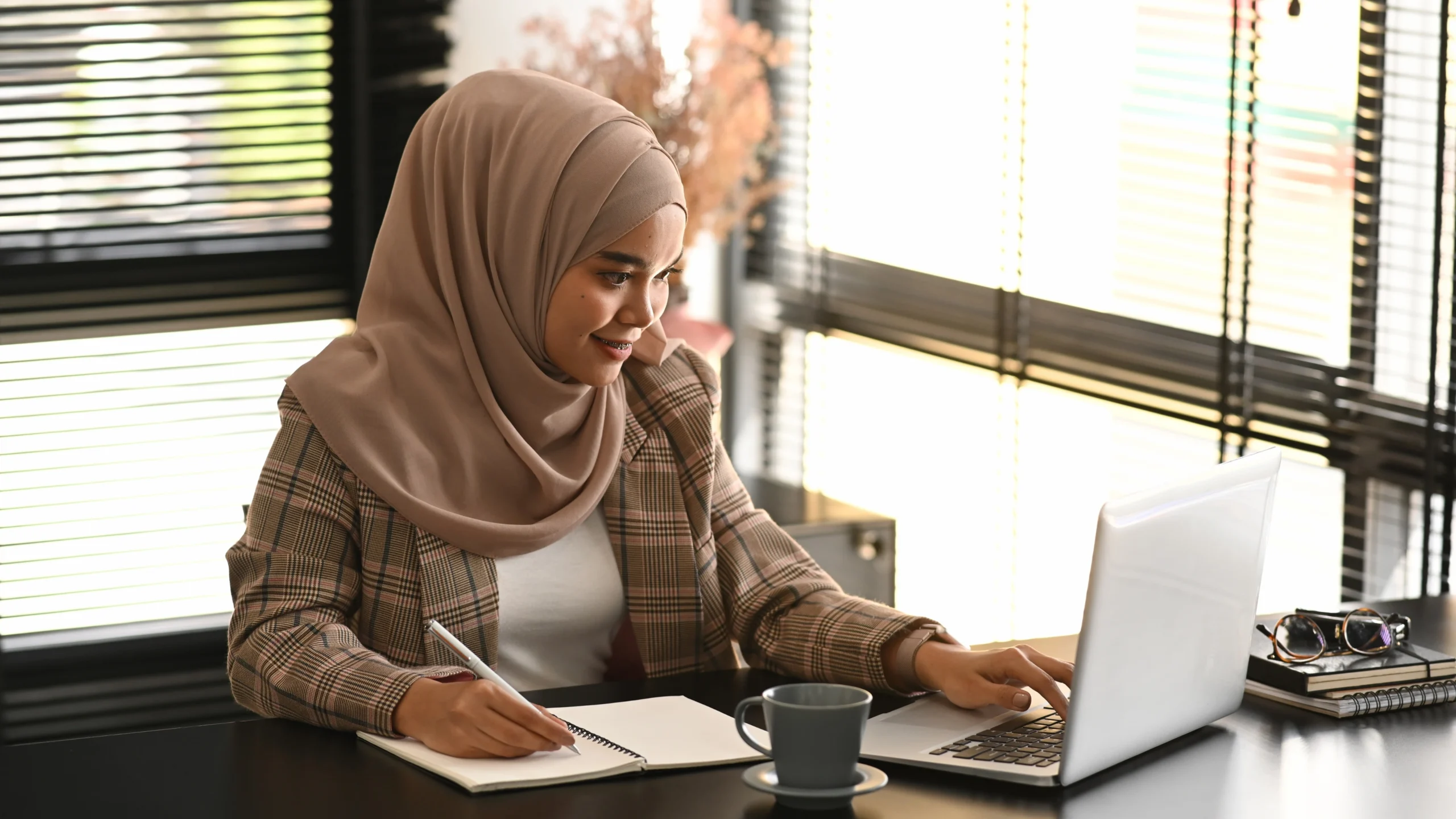 a woman in her office, writing something down while using her laptop