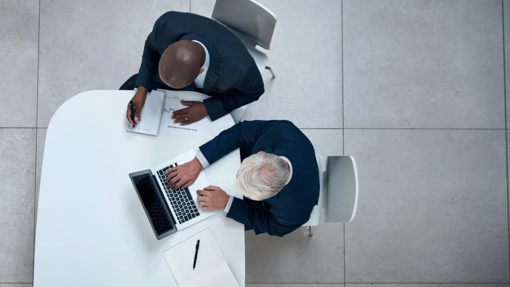 two people sitting at a desk and looking over the same laptop