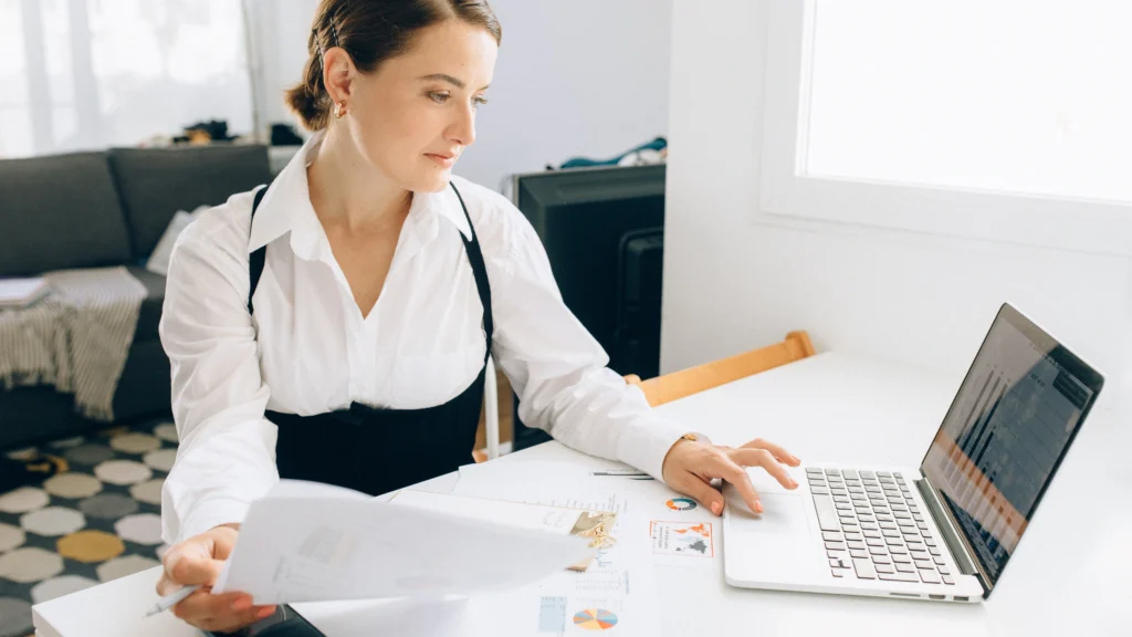 a woman in her work space using a laptop and holding a document