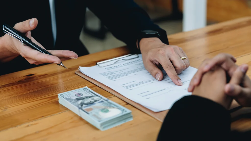 a stack of several 100 dollar bills on the desk while two men talk about business