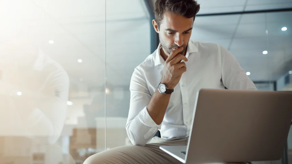 a man sitting near a window in his workplace struggling to choose between two options on his laptop